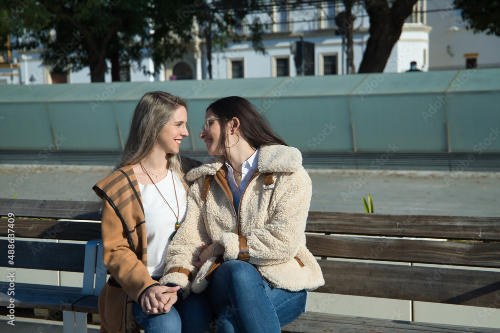 Real couple of young women, holding hands, sitting on a bench next to each other, looking into each other's eyes in an affectionate attitude. Concept lgtbiq+, lesbians, in love, inclusion.