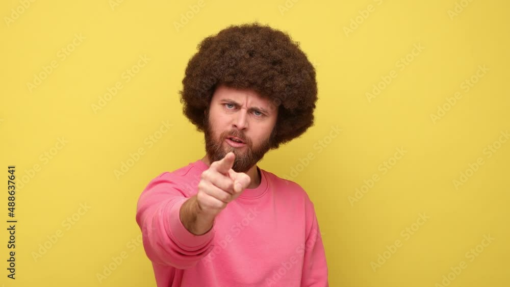 Bearded man with Afro hairstyle pointing at camera with angry expression, accusing blaming, indicating at you, making choice, wearing pink sweatshirt. Indoor studio shot isolated on yellow background. Bearded man with Afro hairstyle pointing at camera with angry expression, accusing blaming, indicating at you, making choice, wearing pink sweatshirt. Indoor studio shot isolated on yellow background.