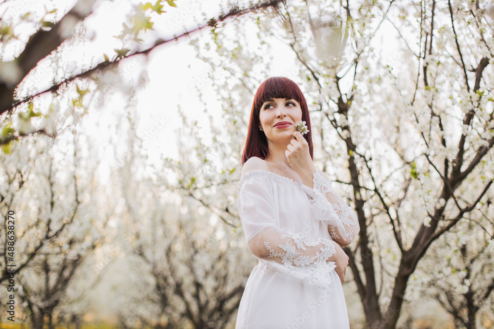 Tender young woman in delicate white dress with bare shoulders posing among spring garden with blooming apple trees. Nature, beauty and sensuality concept. Tender young woman in delicate white dress with bare shoulders posing among spring garden with blooming apple trees. Nature, beauty and sensuality concept.