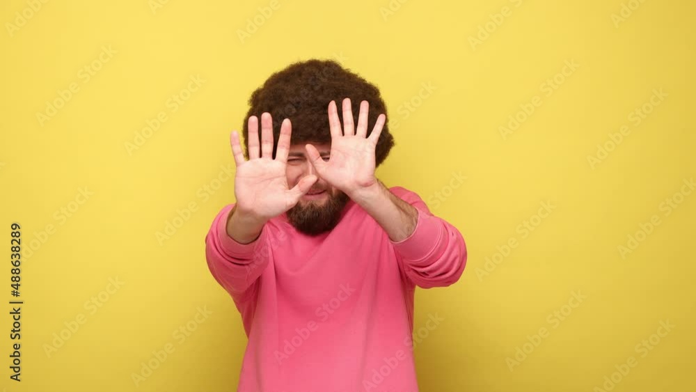 Scared bearded man with Afro hairstyle with raised arms, sees something awful, afraids, stop gesture, wearing pink sweatshirt. Indoor studio shot isolated on yellow background. Scared bearded man with Afro hairstyle with raised arms, sees something awful, afraids, stop gesture, wearing pink sweatshirt. Indoor studio shot isolated on yellow background.
