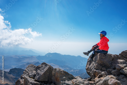 Tableau sur toile A young cheerful girl in a helmet, sits on the top of the mountain higher than c