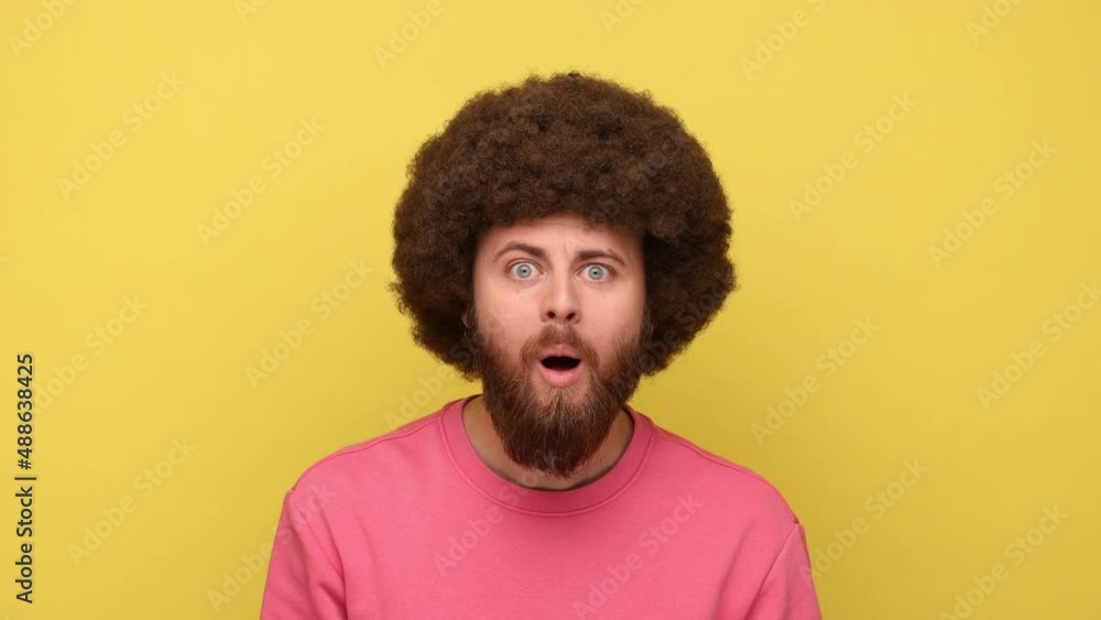 Oh my god, wow. Astonished bearded man with Afro hairstyle looking at camera with mouth open in amazement, expressing shock, wearing pink sweatshirt. Indoor studio shot isolated on yellow background. Oh my god, wow. Astonished bearded man with Afro hairstyle looking at camera with mouth open in amazement, expressing shock, wearing pink sweatshirt. Indoor studio shot isolated on yellow background.