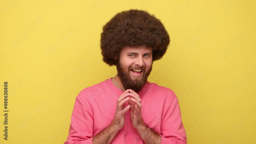 Bearded man with Afro hairstyle thinking devious plan with cunning face expression, having slyly idea to prank, wearing pink sweatshirt. Indoor studio shot isolated on yellow background. Bearded man with Afro hairstyle thinking devious plan with cunning face expression, having slyly idea to prank, wearing pink sweatshirt. Indoor studio shot isolated on yellow background.