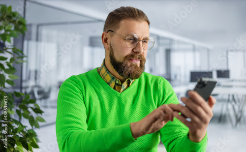 Focused bearded man in bright green sweater and eyeglasses using modern smartphone while working at bright office. Concept of people, business and gadgets.