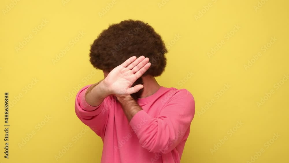 Bearded man with Afro hairstyle standing rising one hand and closing eyes with another, nightmare, eyesight problems, wearing pink sweatshirt. Indoor studio shot isolated on yellow background. Bearded man with Afro hairstyle standing rising one hand and closing eyes with another, nightmare, eyesight problems, wearing pink sweatshirt. Indoor studio shot isolated on yellow background.