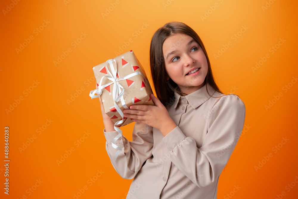 Cute teen girl holding a Christmas gift box against orange background.