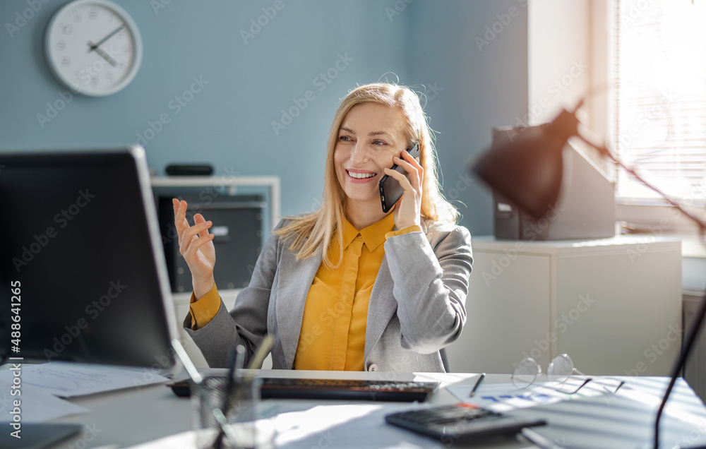 Obraz premium Positive mature woman in stylish formal wear talking on mobile phone while sitting at office desk with modern pc. Working process of caucasian business lady.