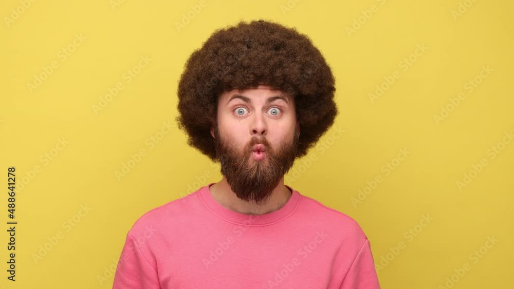 Man with Afro hairstyle making fish face with pout lips and funny childish expression looking at camera with toothy smile, wearing pink sweatshirt. Indoor studio shot isolated on yellow background. Man with Afro hairstyle making fish face with pout lips and funny childish expression looking at camera with toothy smile, wearing pink sweatshirt. Indoor studio shot isolated on yellow background.