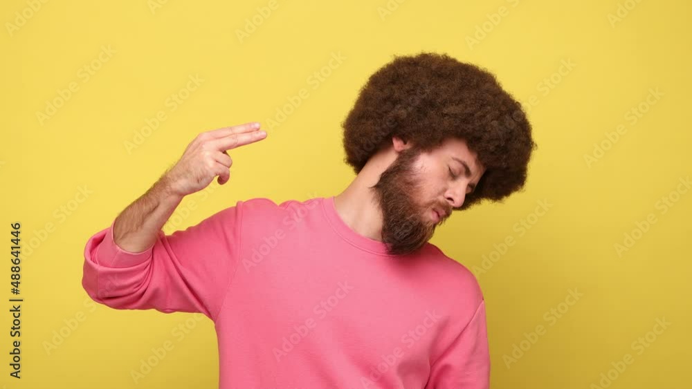 Bearded man with Afro hairstyle looking at camera, making finger gun pointed to head, shooting himself with hand pistol, wearing pink sweatshirt. Indoor studio shot isolated on yellow background. Bearded man with Afro hairstyle looking at camera, making finger gun pointed to head, shooting himself with hand pistol, wearing pink sweatshirt. Indoor studio shot isolated on yellow background.