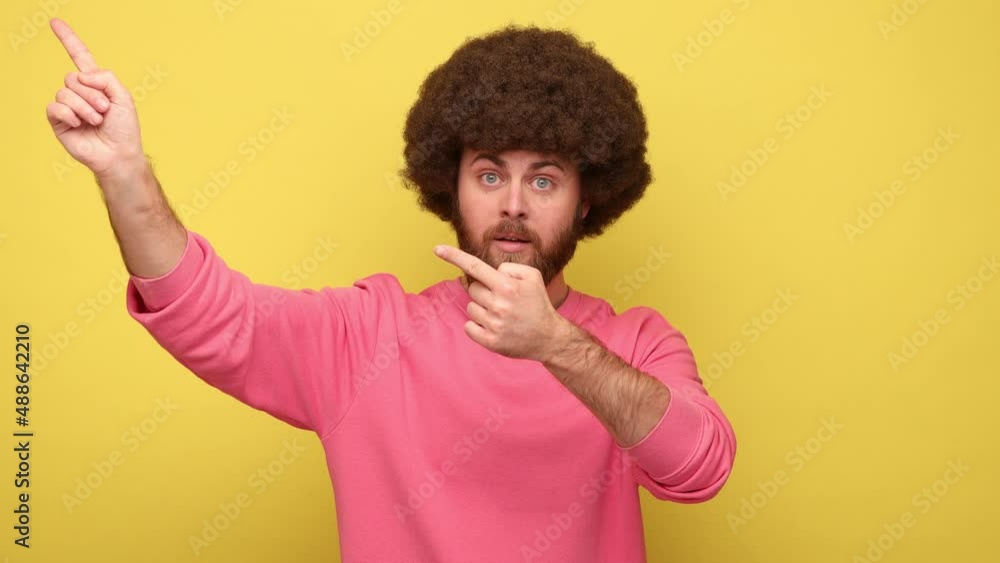 Bearded man with Afro hairstyle looking at camera and pointing finger another way, ordering to leave with bossy expression, wearing pink sweatshirt. Indoor studio shot isolated on yellow background. Bearded man with Afro hairstyle looking at camera and pointing finger another way, ordering to leave with bossy expression, wearing pink sweatshirt. Indoor studio shot isolated on yellow background.