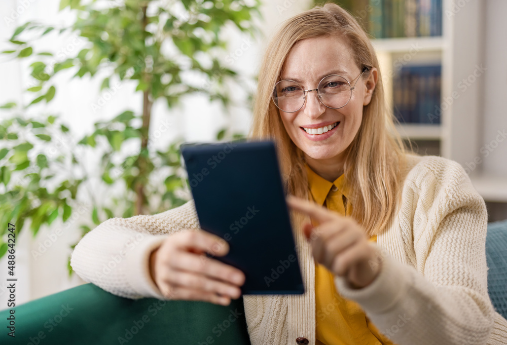 Happy caucasian woman in casual wear and eyegasses using digital tablet whiel resring on couch. Pleasant lady surfing internet during free time at home. Happy caucasian woman in casual wear and eyegasses using digital tablet whiel resring on couch. Pleasant lady surfing internet during free time at home.