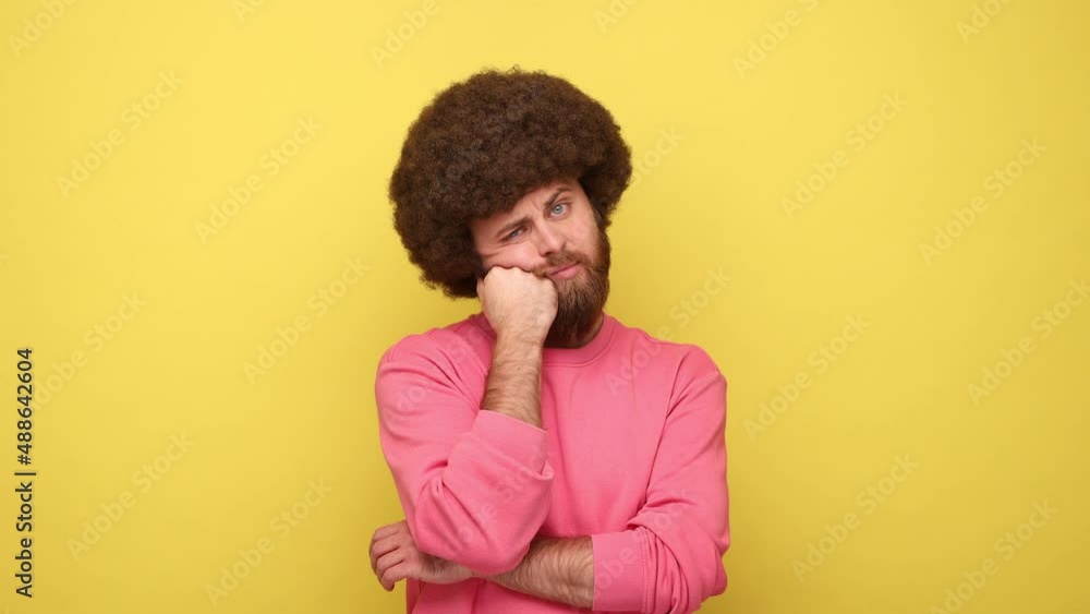 Bearded bored man with Afro hairstyle leaning head on hand laziness and apathy, procrastination, looks sleepy, wearing pink sweatshirt. Indoor studio shot isolated on yellow background. Bearded bored man with Afro hairstyle leaning head on hand laziness and apathy, procrastination, looks sleepy, wearing pink sweatshirt. Indoor studio shot isolated on yellow background.
