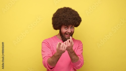 Bearded man with Afro hairstyle making beckoning gesture, inviting for date and looking alluring, flirting to camera, wearing pink sweatshirt. Indoor studio shot isolated on yellow background.