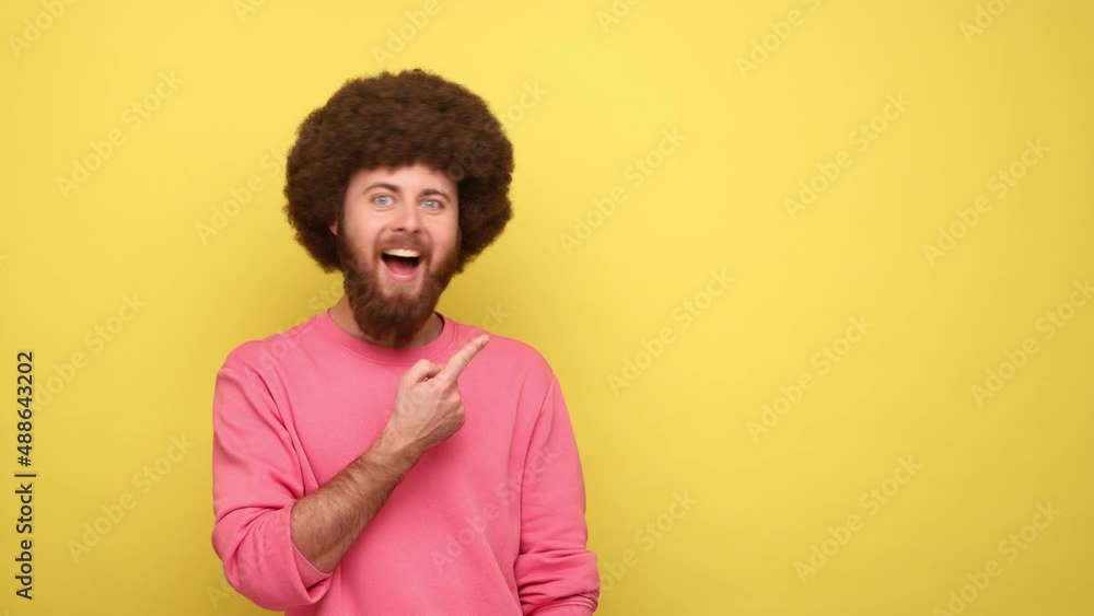 Happy optimistic man with Afro hairstyle presenting copy space on palm, showing empty place for commercial text or goods, wearing pink sweatshirt. Indoor studio shot isolated on yellow background. Happy optimistic man with Afro hairstyle presenting copy space on palm, showing empty place for commercial text or goods, wearing pink sweatshirt. Indoor studio shot isolated on yellow background.