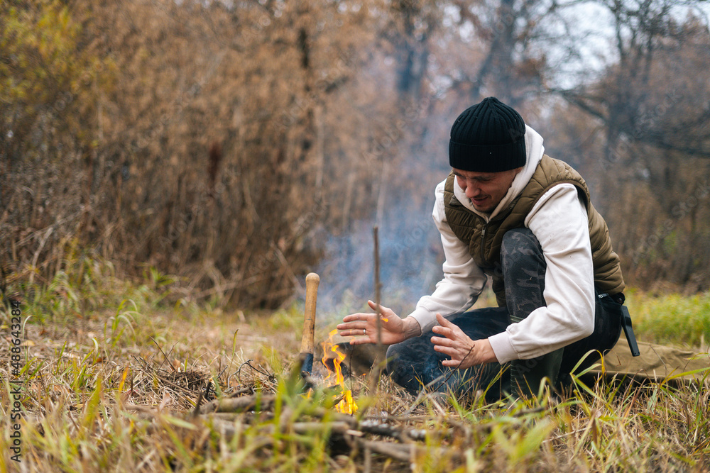 Portrait of frozen tourist male wearing warm clothes warming hands over fire at outdoors on overcast cold day. Concept of scout, research, travel and survival in nature. Portrait of frozen tourist male wearing warm clothes warming hands over fire at outdoors on overcast cold day. Concept of scout, research, travel and survival in nature.