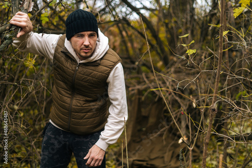 Tired lost hiker male wearing warm clothes wading through thickets in forest in cold overcast day, looking at camera. Concept of bushcraft, camping and survival in nature.