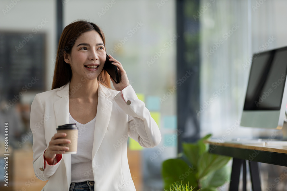 Beautiful Asian woman talking on the phone and holding a hot cup of coffee in her office, she is a businesswoman who runs a startup company with her partner. Startup company concept. Beautiful Asian woman talking on the phone and holding a hot cup of coffee in her office, she is a businesswoman who runs a startup company with her partner. Startup company concept.