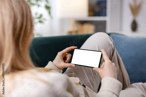 Back view of woman with blond hair resting on couch and holding modern smartphone in horizontal position. Focus on white empty screen of mobile.
