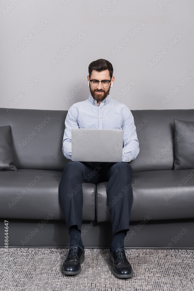 happy man in eyeglasses and earphones using laptop while sitting on sofa in hotel happy man in eyeglasses and earphones using laptop while sitting on sofa in hotel