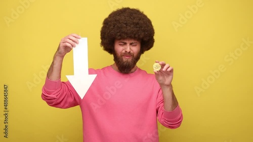 Bearded hipster man with Afro hairstyle showing gold bitcoin and arrow pointing down, downgrade of electronic currency, wearing pink sweatshirt. Indoor studio shot isolated on yellow background. photo