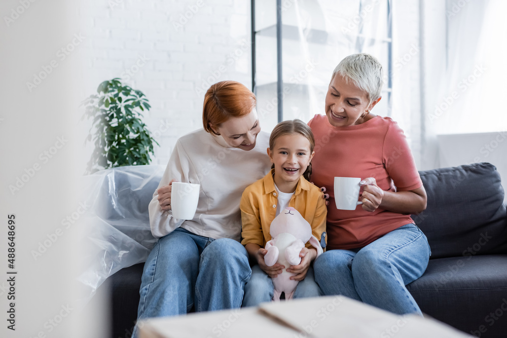 cheerful girl with toy bunny looking at camera near same sex mothers sitting with tea cups in new home cheerful girl with toy bunny looking at camera near same sex mothers sitting with tea cups in new home