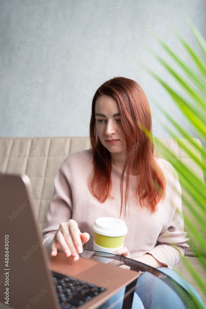 Redhead young woman sitting on sofa with laptop and eco cup. Redhead young woman sitting on sofa with laptop and eco cup.