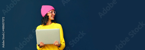 Portrait of modern happy smiling young woman with laptop looking away wearing colorful clothes on dark blue background, banner blank copy space for advertising text