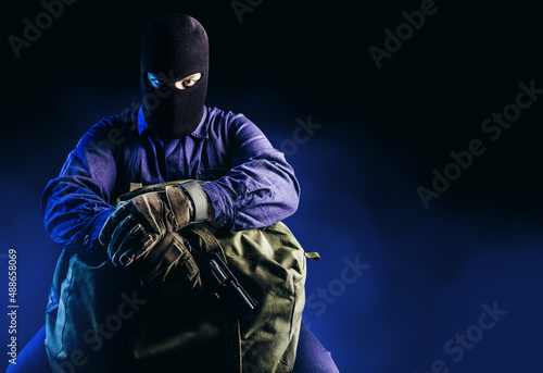Fototapeta Photo of robber in mask, overalls, gloves, gun sitting and holding bag on dark blue background