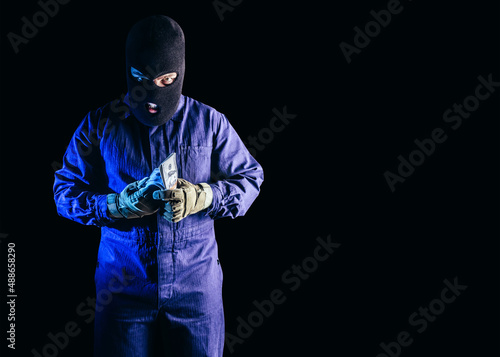 Photo of robber in mask, overalls, gloves standing and counting money on black background.