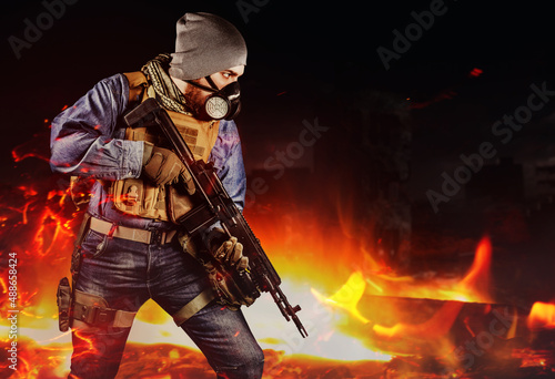 Photo of urban soldier in tactical military outfit and gas mask standing with rifle and gas mask on dark destroyed and burning city background.