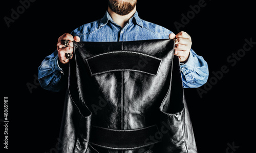 Photo of biker man in jeans shirt holding leather vest with patch colors on black background.