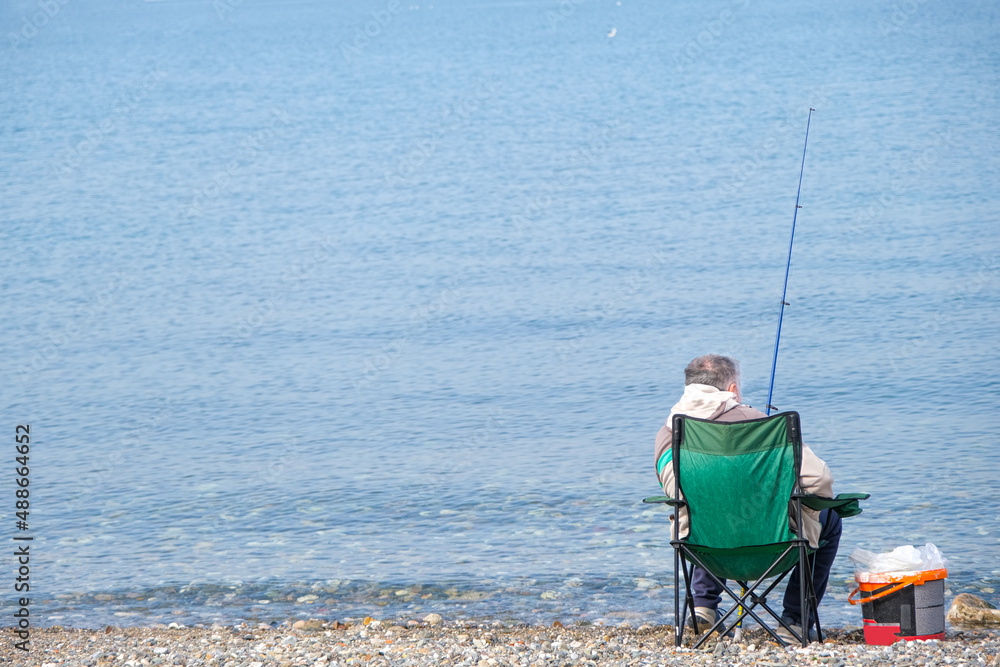 Man fishing on beach, one single male sitting on chair with fishing rod on coast with calm blue sea background. Beautiful minimalist concept with area for ad or advertisement text. Man fishing on beach, one single male sitting on chair with fishing rod on coast with calm blue sea background. Beautiful minimalist concept with area for ad or advertisement text.