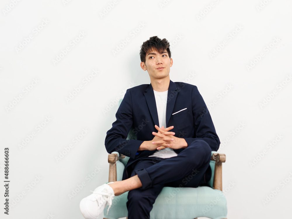 Portrait of handsome Chinese young man in dark blue leisure suit sitting in armchair and posing against white wall background. Hands clasped on crossed leg and looking away seriously. Portrait of handsome Chinese young man in dark blue leisure suit sitting in armchair and posing against white wall background. Hands clasped on crossed leg and looking away seriously.