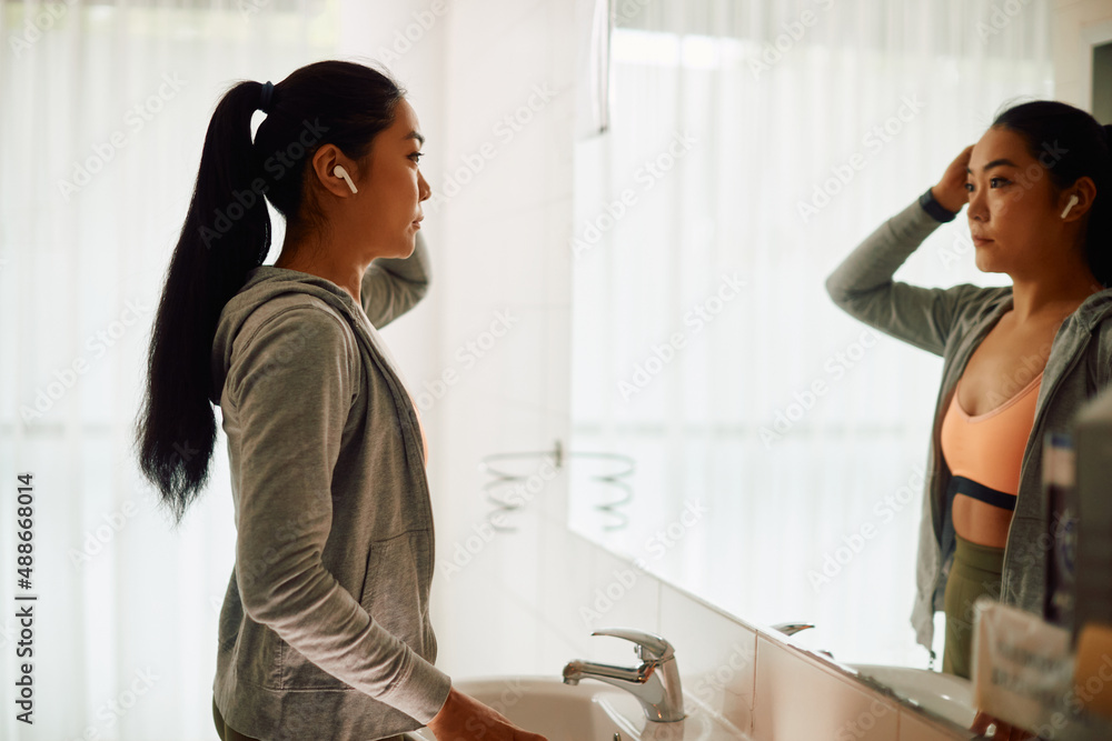 Obraz premium Young Asian athlete looks herself in mirror at gym's bathroom.