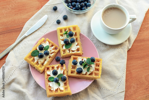 Waffles with cream cheese and blueberries on a pink plate, coffee cup on wooden table. Breakfast concept. Top view, flt lay