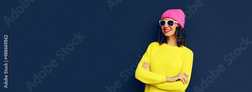 Portrait of stylish smiling young woman posing wearing colorful clothes, yellow knitted sweater and pink hat on black blue background, banner blank copy space for advertising text © rohappy
