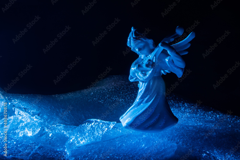 Photo of blue toned ice angel miniature playing the lute and standing on cracked ice surface on dark backdrop.