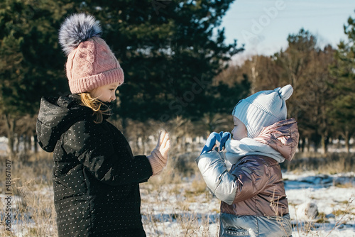 Two sisters on a walk in a winter park stand opposite each other in warm clothes, in mittens and gloves and play patties.