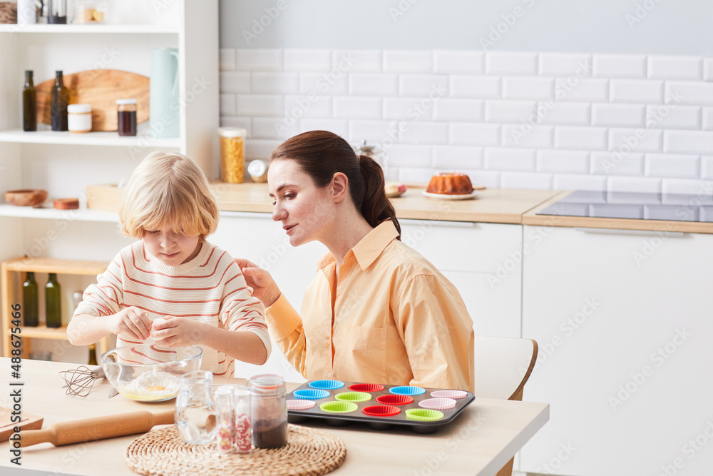 Fototapeta premium Portrait of young woman baking cupcakes with cute little boy helping mom in kitchen, copy space