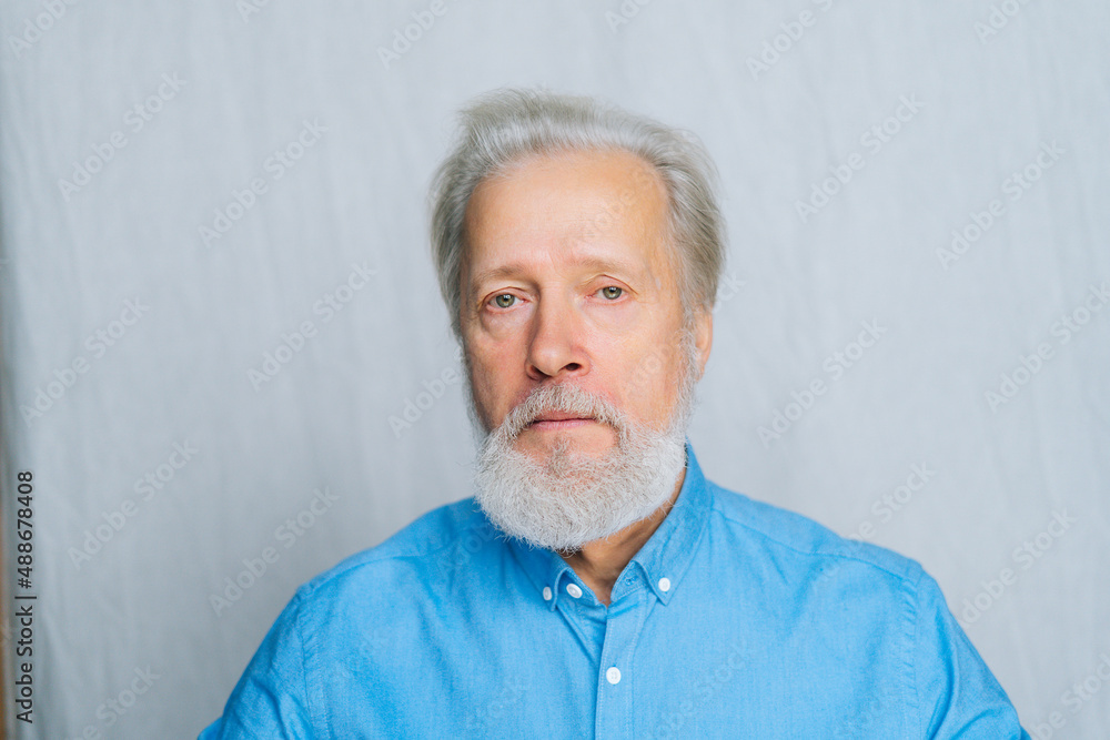 Close-up portrait of serious gray-haired senior man unemotionally looking at camera on white isolated background. Front view of handsome bearded mature aged gentleman posing in studio. Close-up portrait of serious gray-haired senior man unemotionally looking at camera on white isolated background. Front view of handsome bearded mature aged gentleman posing in studio.