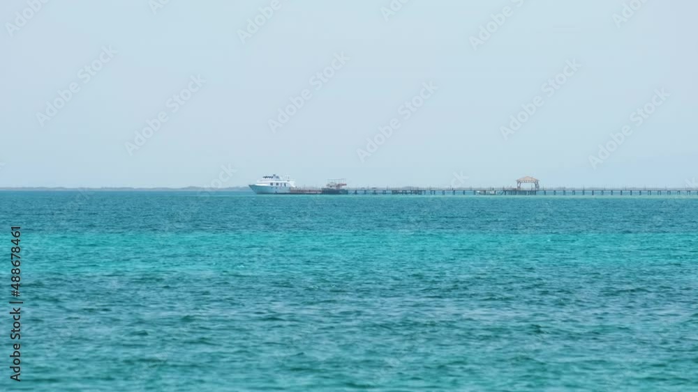 Seascape with ripple surface of blue sea water with distant ship at the end of long pier floating on calm waves Seascape with ripple surface of blue sea water with distant ship at the end of long pier floating on calm waves