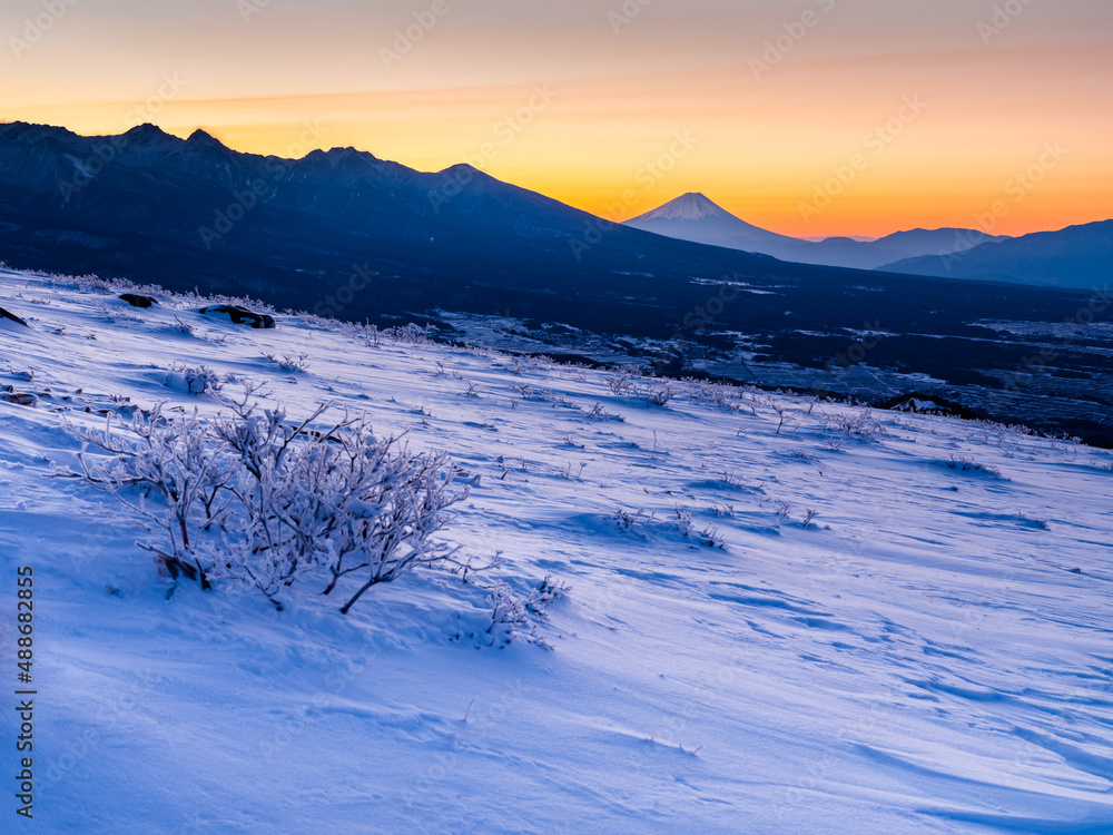 車山より冬山の早朝で雪面も空も一面に朝焼けのオレンジ色に染まり、山並みの隅には遠くの富士山が見える。
