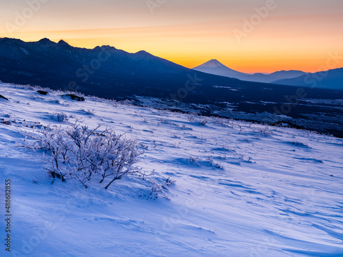 車山より冬山の早朝で雪面も空も一面に朝焼けのオレンジ色に染まり、山並みの隅には遠くの富士山が見える。