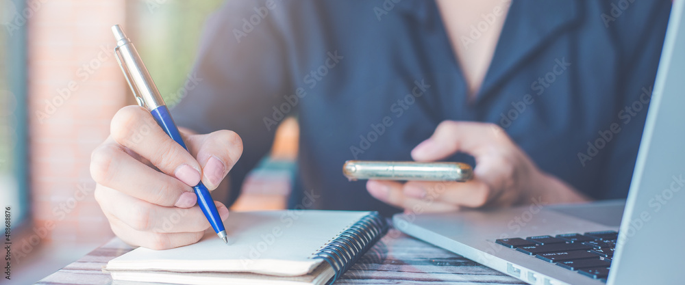 Woman hand writing on a notepad with a pen and use a smartphone in the office.