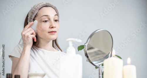 Woman is looking in the mirror and doing a massage with a gouache scraper. Candles and an aroma lamp with essential oils on the table. Concept of facial care, aromatherapy, morning and evening rituals