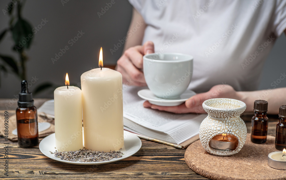 Woman is reading a book and drinking tea in atmosphere of harmony and relaxation. Aroma lamp with essential oils and burning candles on the wooden table