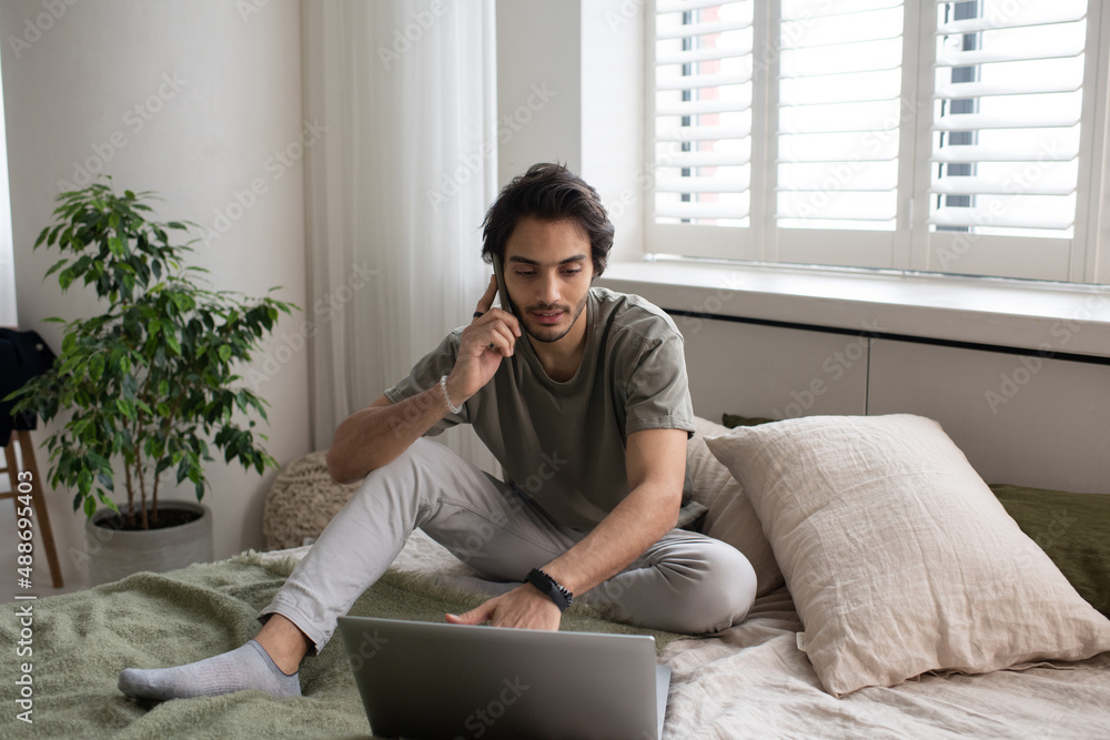 Young contemporary busy employee or freelancer talking on smartphone and typing on laptop keyboard while sitting on bed at home