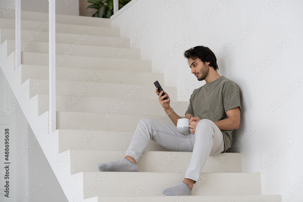 Young modern manager in casualwear sitting on white staircase, having drink and scrolling in mobile phone while working from home