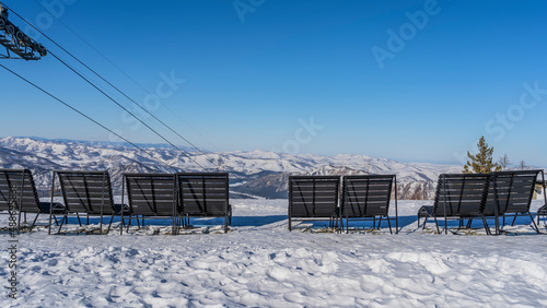 Wooden benches stand on a snowy plateau. Ahead is a mountain range. The cable car ropes are visible. Altai Republic. Manzherok