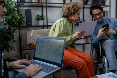 Mature economist analyzing data on laptop screen against two young intercultural coworkers discussing financial document